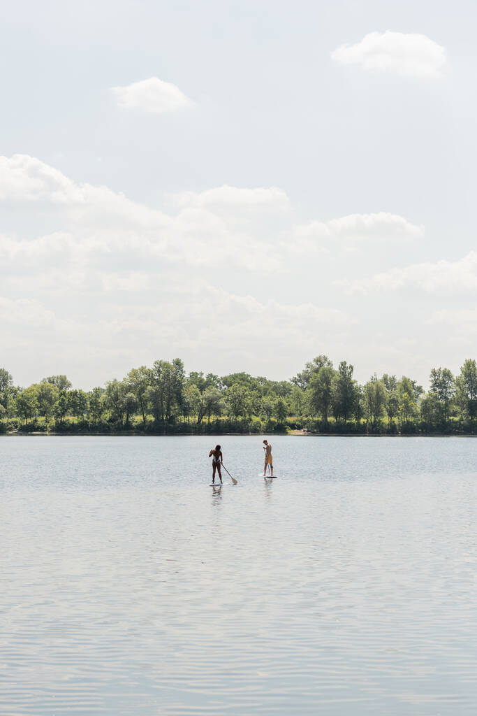pohled z dálky sportovní mezirasový pár plachtění na sup desky při trávení víkendu na jezeře se zeleným malebným pobřežím pod oblačným nebem, venkovní aktivity, letní vibrace - Fotografie, Obrázek