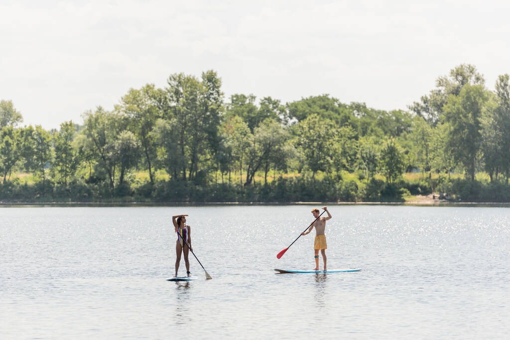 по всей длине африканская американская женщина плавает на доске и глядя в сторону рядом молодой и спортивный мужчина в плавках на живописном озере с зелеными деревьями на берегу - Фото, изображение