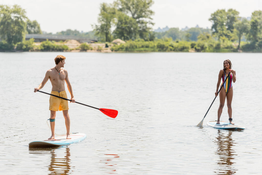 longitud completa de la pareja deportiva interracial sosteniendo paletas y sonriéndose entre sí mientras navegan en tablas de sup en el río durante la recreación acuática en el fin de semana de verano - Foto, imagen
