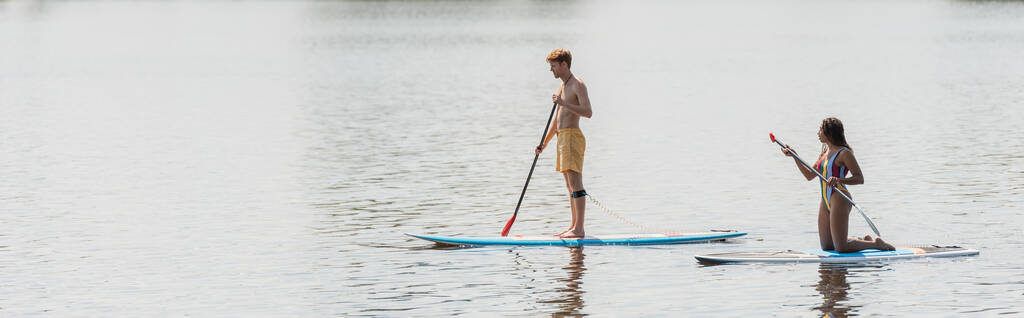 pełna długość ruda i sportowy mężczyzna żeglarstwo na pokładzie w pobliżu Afryki amerykańskiej kobiety w paski strój kąpielowy na spokojnej rzeki w letni weekend, sztandar - Zdjęcie, obraz