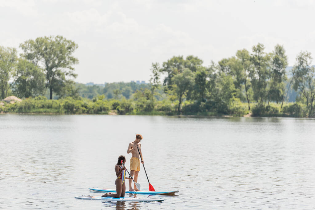 active multiethnic couple in colorful swimwear sailing on sup boards with paddles while spending summer vacation day on river with green trees on scenic bank - Photo, Image