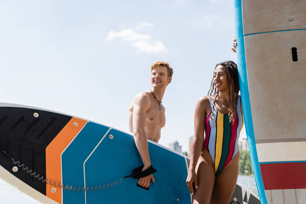 carefree redhead man and sportive african american woman in colorful swimsuit holding sup boards, smiling and looking away during summer weekend near river - Photo, Image