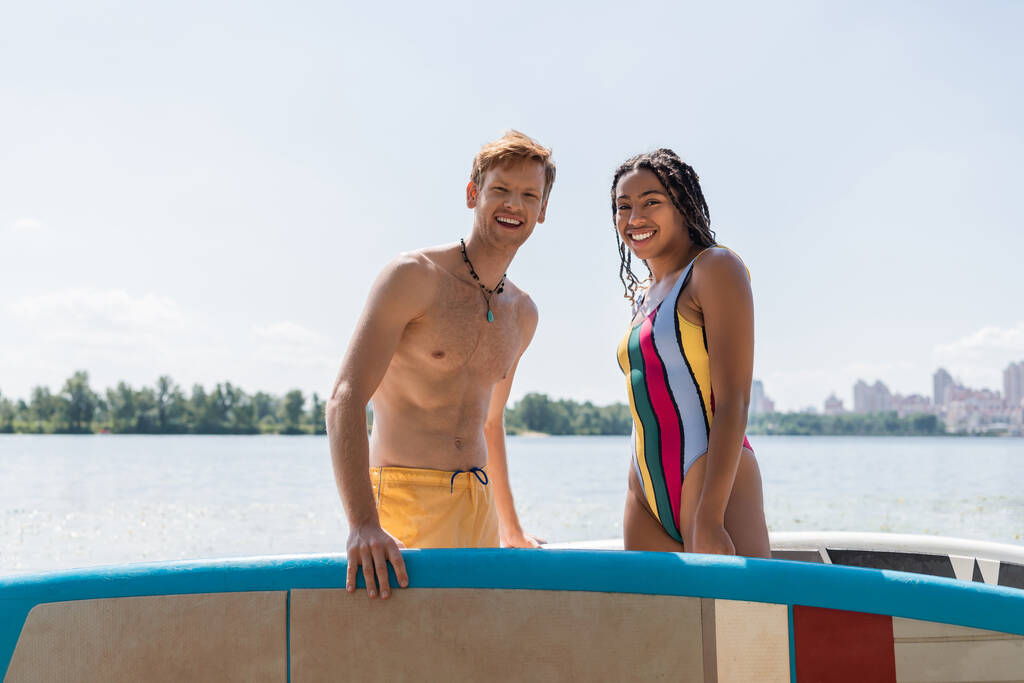 séduisante femme afro-américaine en maillot de bain rayé et jeune homme rousse en short de bain jaune regardant la caméra près des planches à soup avec lac et paysage urbain en arrière-plan - Photo, image