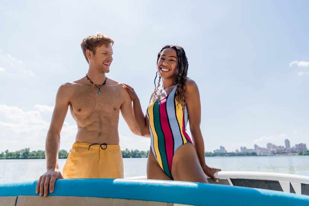 cheerful and enchanting african american woman in colorful swimsuit looking at camera near young and redhead man and sup boards under blue sky outdoors - Photo, Image