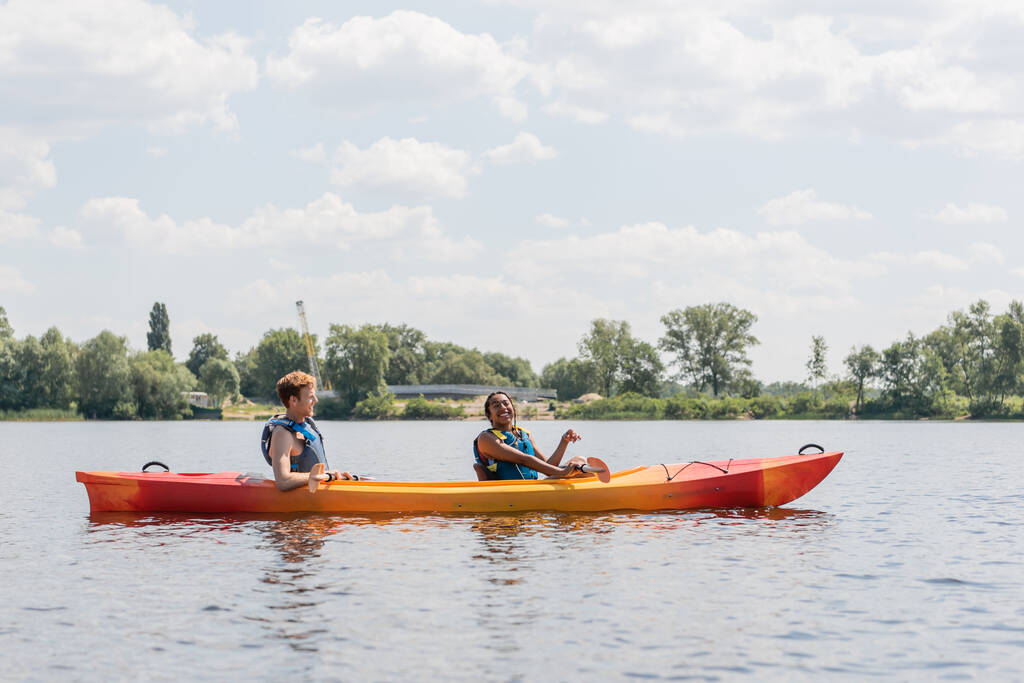 vista lateral de la alegre mujer afroamericana y el joven pelirrojo en chalecos salvavidas navegando en kayak deportivo en el río con la orilla verde bajo el cielo azul y nublado - Foto, imagen