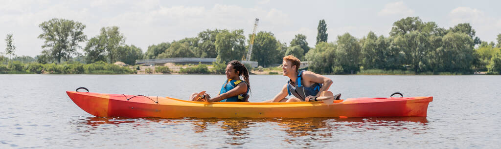 Seitenansicht des sportlichen und gemischtrassigen Paares in Schwimmwesten, die im Kajak im Sommer Wassererholung auf dem Fluss mit grünem malerischem Ufer segeln, Banner - Foto, Bild