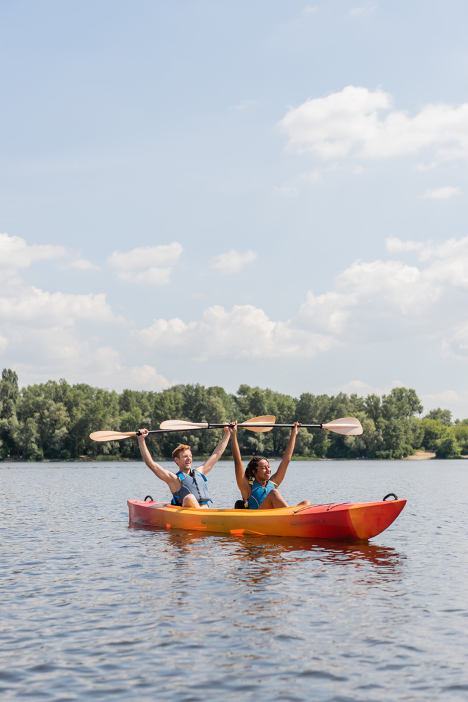 couple of cheerful interracial friends in life vests holding paddles in raised hands while sitting in kayak on picturesque lake under blue cloudy sky with green riverside on background - Photo, Image