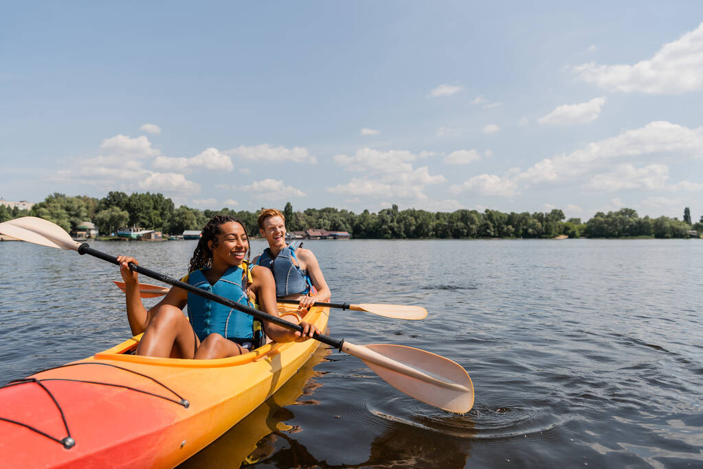 carefree and charming african american woman and young redhead man in life vests sailing in sportive kayak on calm water surface under blue sky with clouds on summer day - Photo, Image