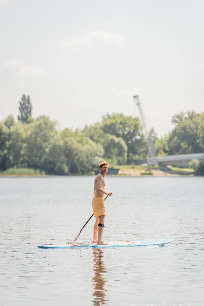 full length of sportive redhead man in yellow swim shorts holding paddle and looking at camera while sailing on picturesque lake on summer weekend day - Photo, Image
