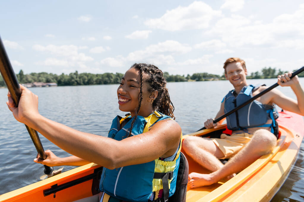 cheerful african american woman in life vest sailing in kayak while spending time on river with young and redhead man paddleboarding on blurred background - Photo, Image