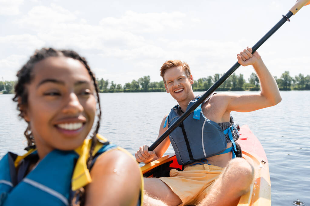 overjoyed redhead man in life vest sitting in kayak and holding paddle while looking at camera near african american woman smiling on blurred foreground - Photo, Image