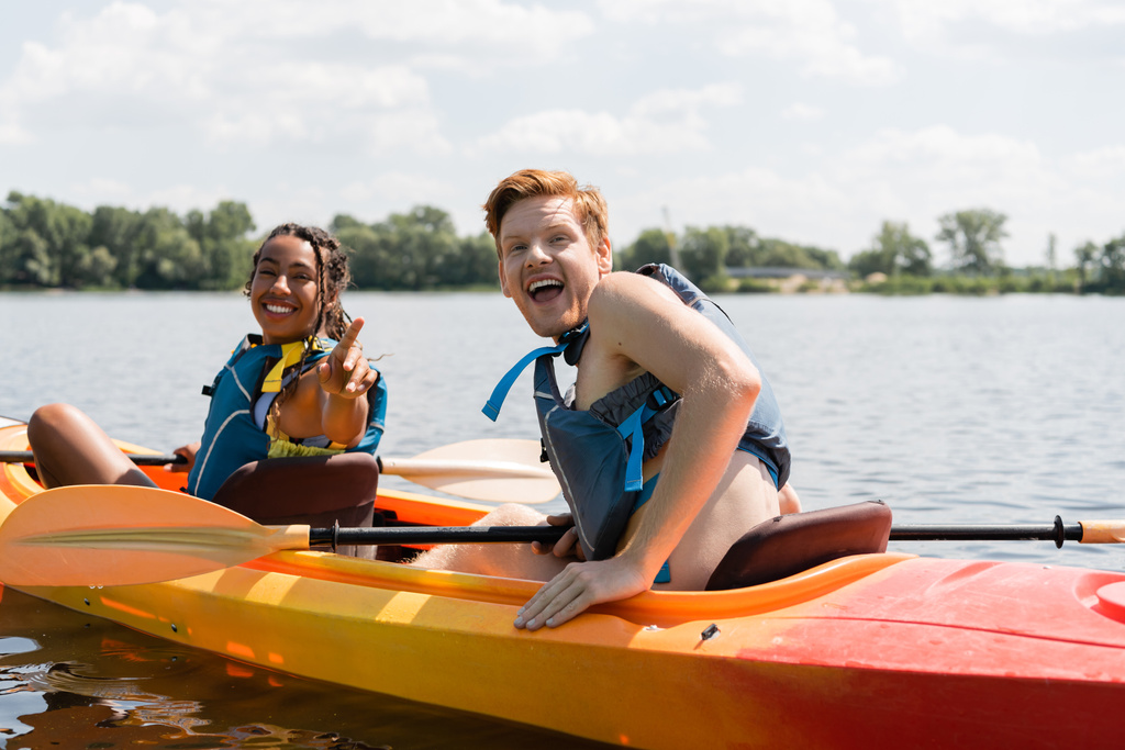 excité rousse homme avec la bouche ouverte regardant caméra près de femme afro-américaine gaie dans la vie gilet pointant du doigt tout en naviguant en kayak sur le lac en été - Photo, image