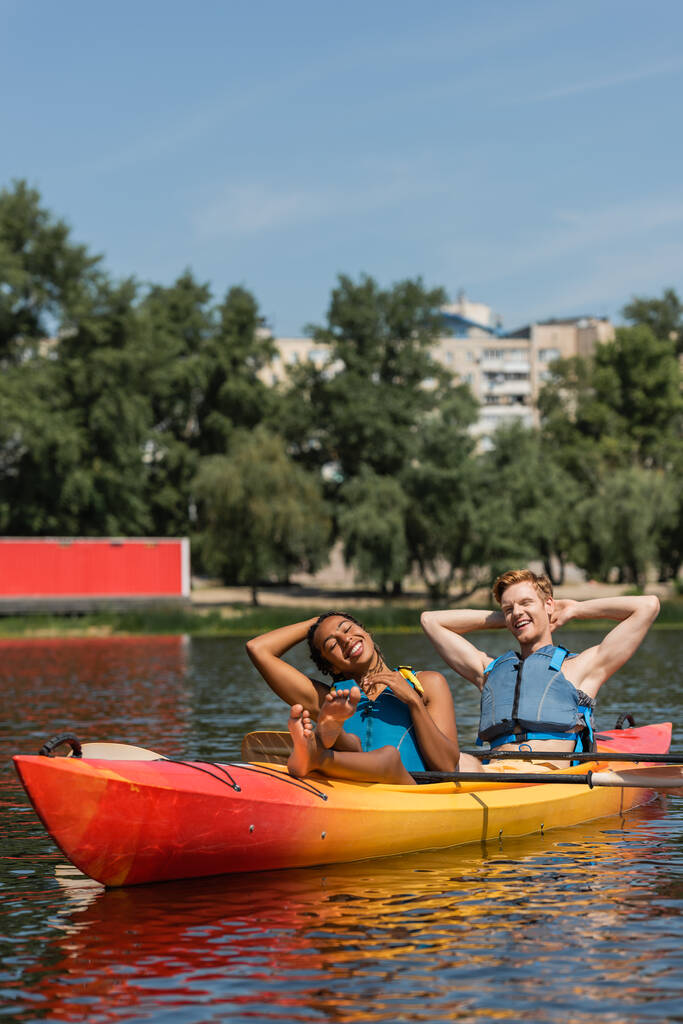 zadowolona i boso afrykańska kobieta i uśmiechnięty rudy mężczyzna w kamizelkach życia relaks z zamkniętymi oczami w sportowym kajaku podczas letniego weekendu nad jeziorem - Zdjęcie, obraz