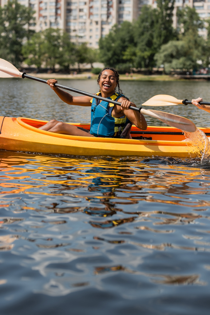 счастливая и спортивная африканская американка в спасательном жилете, держащая весло во время плавания на байдарке с юным другом во время летних выходных на озере в городе - Фото, изображение