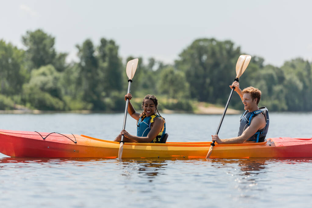 alegre y activa pareja interracial en chalecos salvavidas remando en kayak deportivo mientras pasa tiempo en el lago con borrosa orilla verde durante las vacaciones de verano - Foto, Imagen