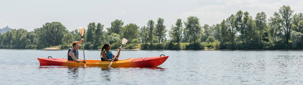 side view of young man and african american woman in life vests sailing in sportive kayak during active summer weekend on picturesque river with green trees on bank, banner - Photo, Image