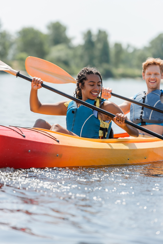 carefree and charming african american woman in life vest holding paddle and sailing in sportive kayak near young redhead man smiling on blurred background - Photo, Image
