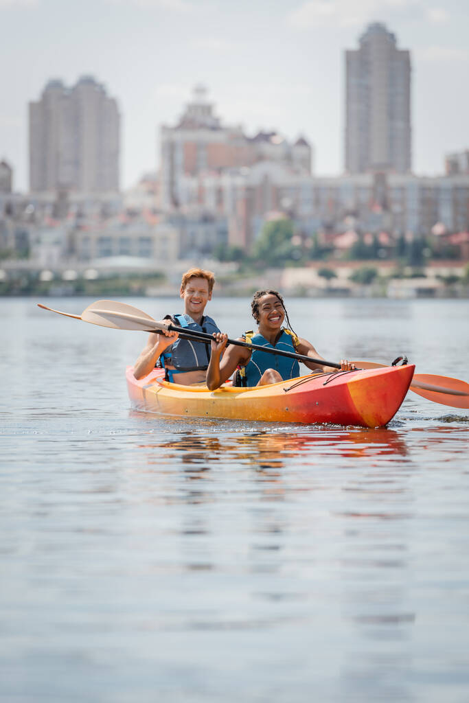 carefree and enchanting african american woman and young redhead man in life vests holding paddles and sailing in sportive kayak on river with scenic cityscape on blurred background - Photo, Image