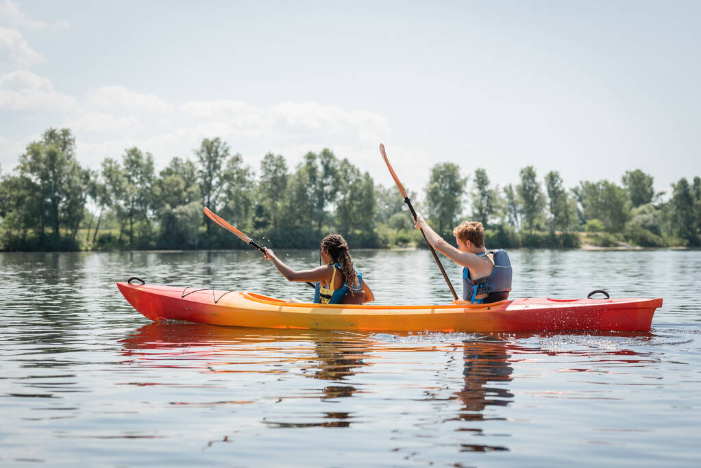 vista lateral de pareja multiétnica activa en chalecos salvavidas sosteniendo paletas mientras navega en kayak deportivo en el río con pintoresca orilla con árboles verdes en verano - Foto, imagen
