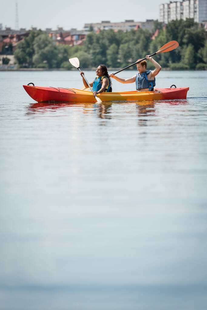 szczęśliwy Afroamerykanka kobieta i młody aktywny mężczyzna w kamizelkach życia spędzać czas na jeziorze miasta i żeglarstwo w sportowym kajaku z wiosłami na zamazanym pierwszym planie - Zdjęcie, obraz