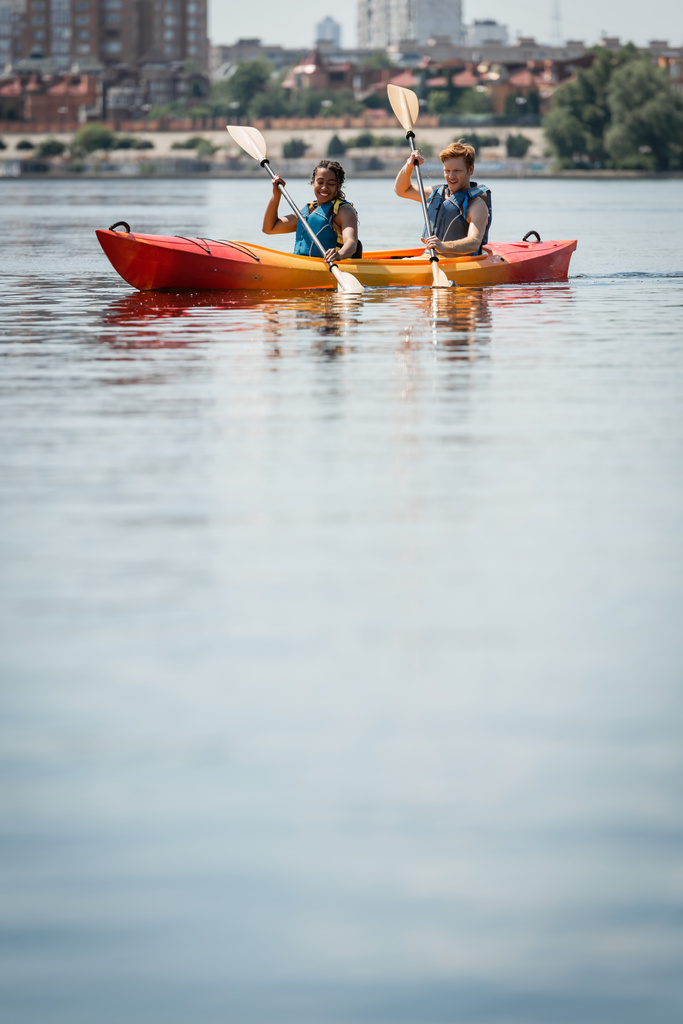 positive and active multiracial couple in life vests holding paddles while sailing in sportive kayak on calm water surface during summer recreation weekend on city lake - Photo, Image
