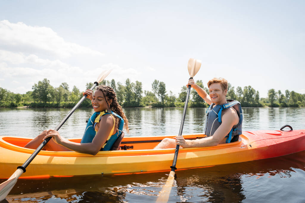 heureuse et charmante femme afro-américaine avec jeune homme rousse tenant des pagaies et naviguant en kayak sportif sur un lac pittoresque pendant un week-end d'été actif - Photo, image