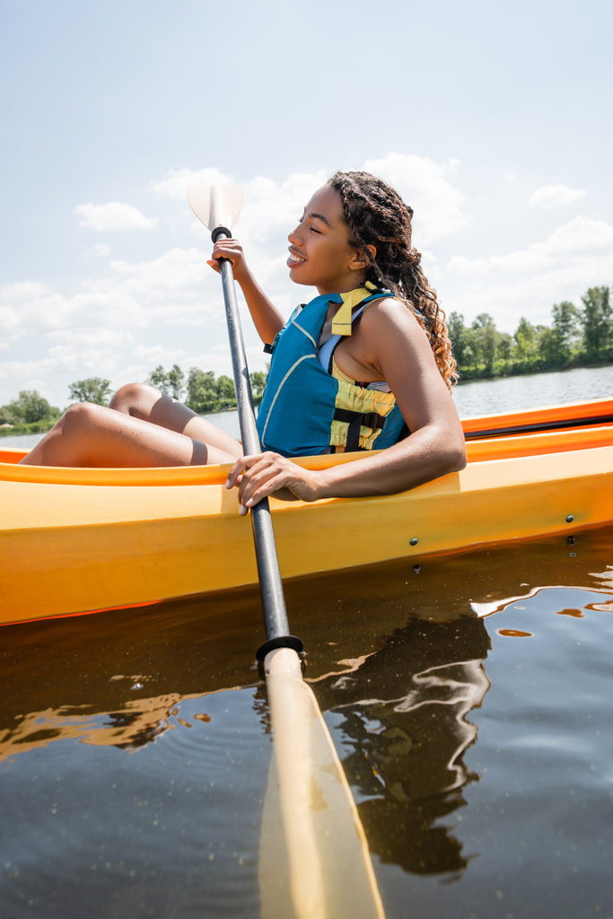 vue latérale de la brune et attrayante femme afro-américaine en gilet de sauvetage tenant la pagaie et la voile en kayak sportif pendant les loisirs aquatiques en été - Photo, image