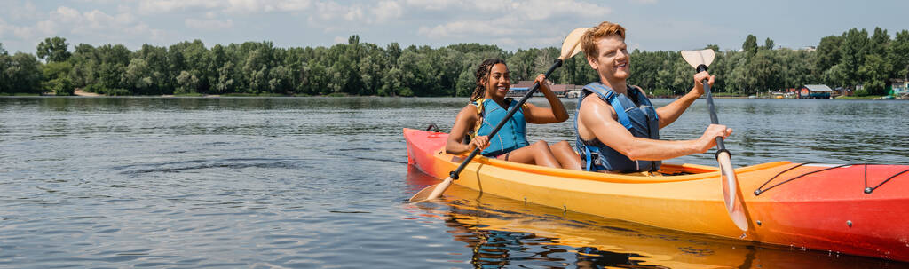 alegre pelirroja hombre y bastante africana americana mujer en chalecos salvavidas remando en kayak deportivo durante el verano recreación en el río con árboles verdes en la orilla, bandera - Foto, Imagen
