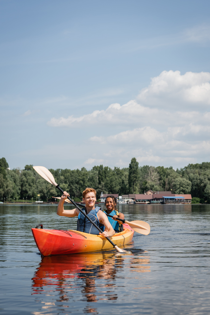 felice e attiva coppia multietnica in giubbotti di salvataggio vela in kayak sportivo con pagaie sul fiume con riva verde sotto il cielo blu con nuvole in estate - Foto, immagini