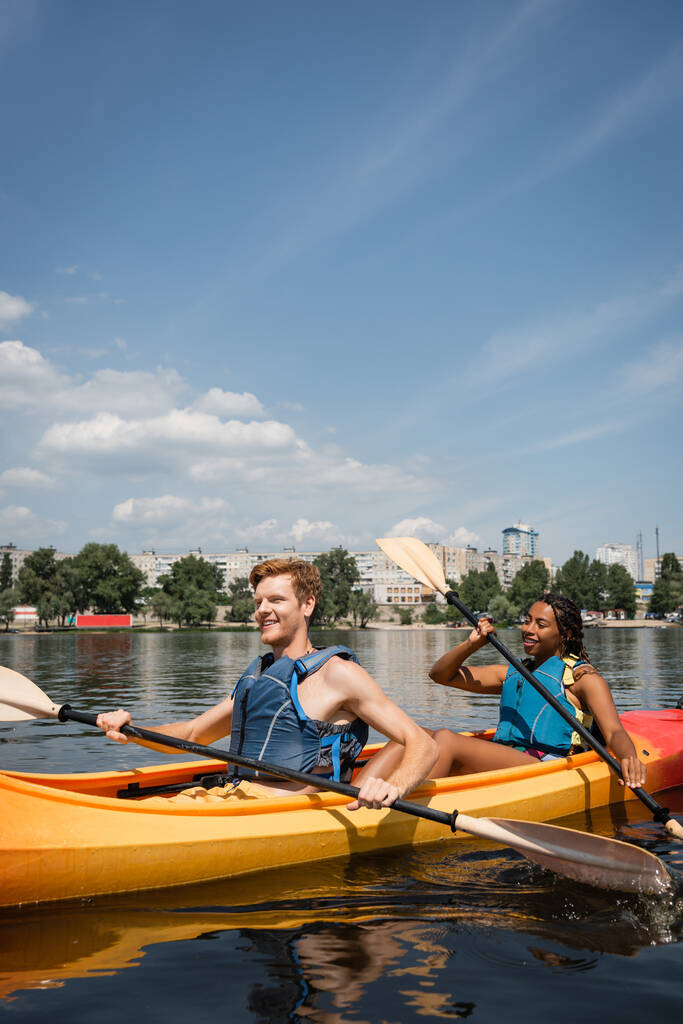 positive and active multiethnic couple in life vests spending recreation weekend by sailing in sportive kayak on city river under blue sky with white clouds - Photo, Image
