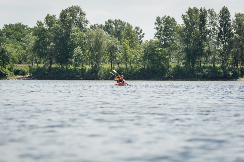 view from afar of active and interracial couple in life vests sailing in sportive kayak during recreation weekend on river with green bank during water recreation in summer - Photo, Image