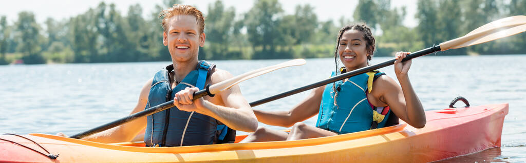 carefree interracial man and woman in life vests holding paddles and smiling at camera while sailing in sportive kayak on lake during summer weekend on blurred background, banner - Photo, Image