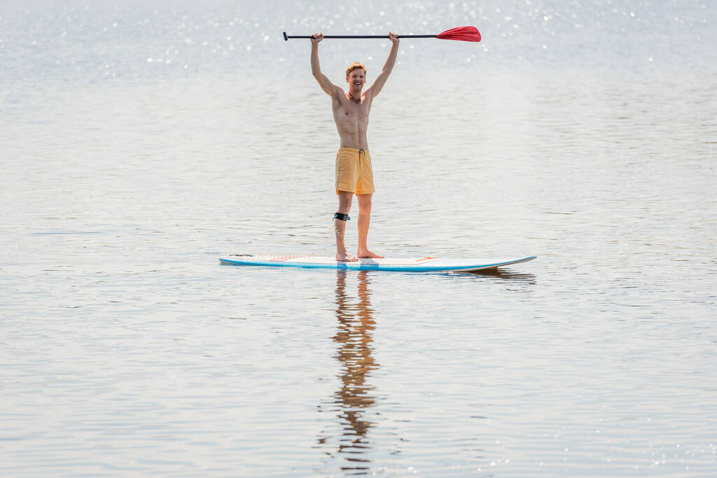 volle Länge eines überglücklichen rothaarigen Mannes in gelben Badehosen mit Paddel in erhobenen Händen, während er am Sommerwochenende auf einem Surfbrett auf einem See mit ruhigem Wasser steht - Foto, Bild