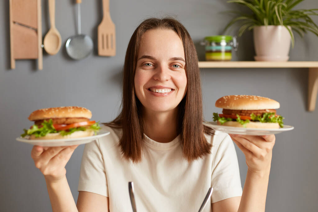 Ανθυγιεινό φαγητό. Όμορφο θετικό κορίτσι χαμογελά στην κάμερα και ποζάρουν με δύο νόστιμα χάμπουργκερ, νεαρή όμορφη γυναίκα φορώντας λευκό t-shirt κρατώντας μεγάλα cheeseburgers. - Φωτογραφία, εικόνα