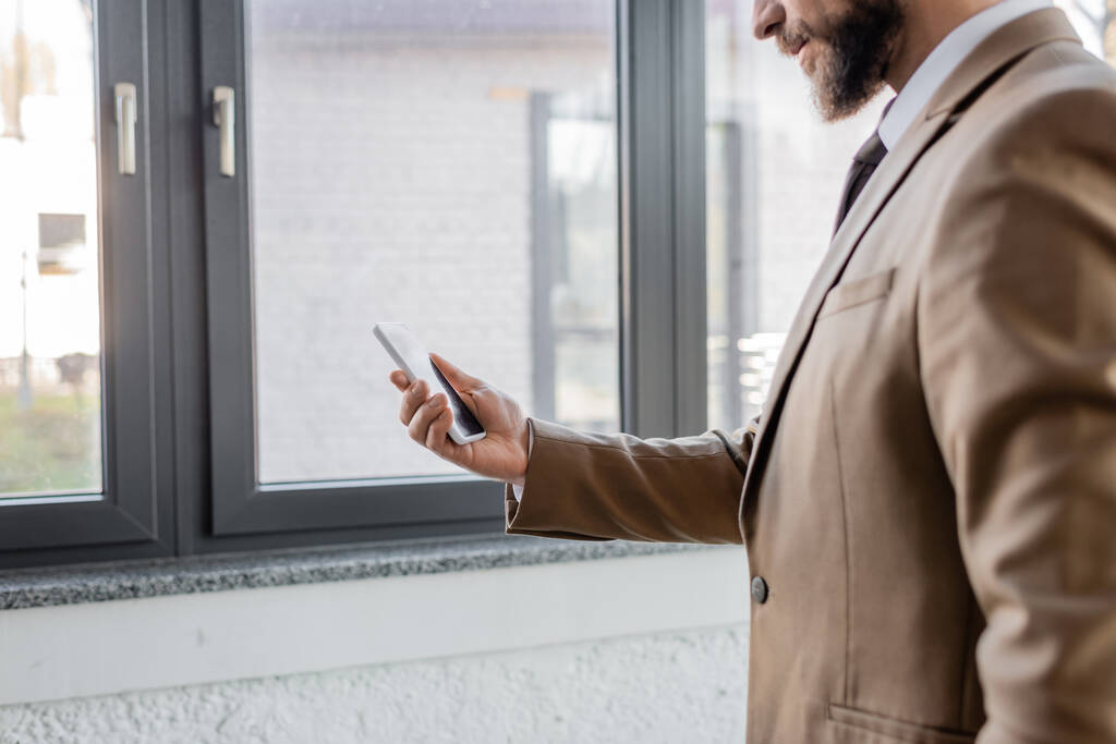 μερική άποψη του γενειοφόρου επιχειρηματία σε μπεζ κομψό σακάκι κρατώντας κινητό τηλέφωνο με λευκή οθόνη, ενώ στέκεται κοντά σε μεγάλα παράθυρα στο σύγχρονο γραφείο - Φωτογραφία, εικόνα