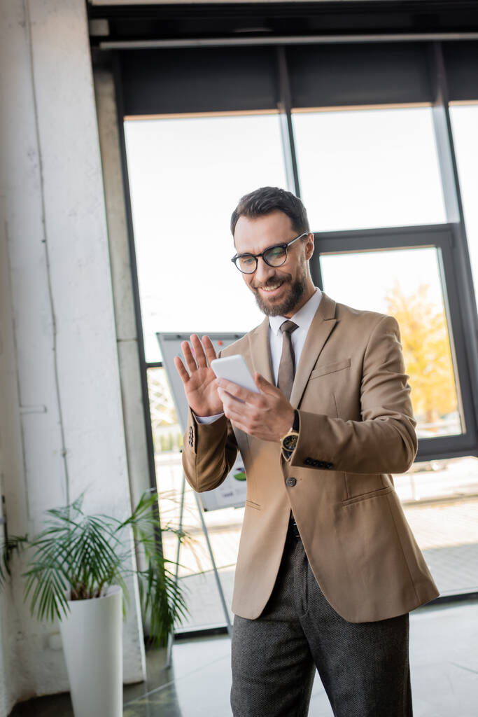 radosny brodaty menedżer w stylowym stroju biznesowym i okularach machających ręką podczas rozmowy wideo na telefon komórkowy w pobliżu flip chart i doniczkowe roślin w nowoczesnym biurze - Zdjęcie, obraz