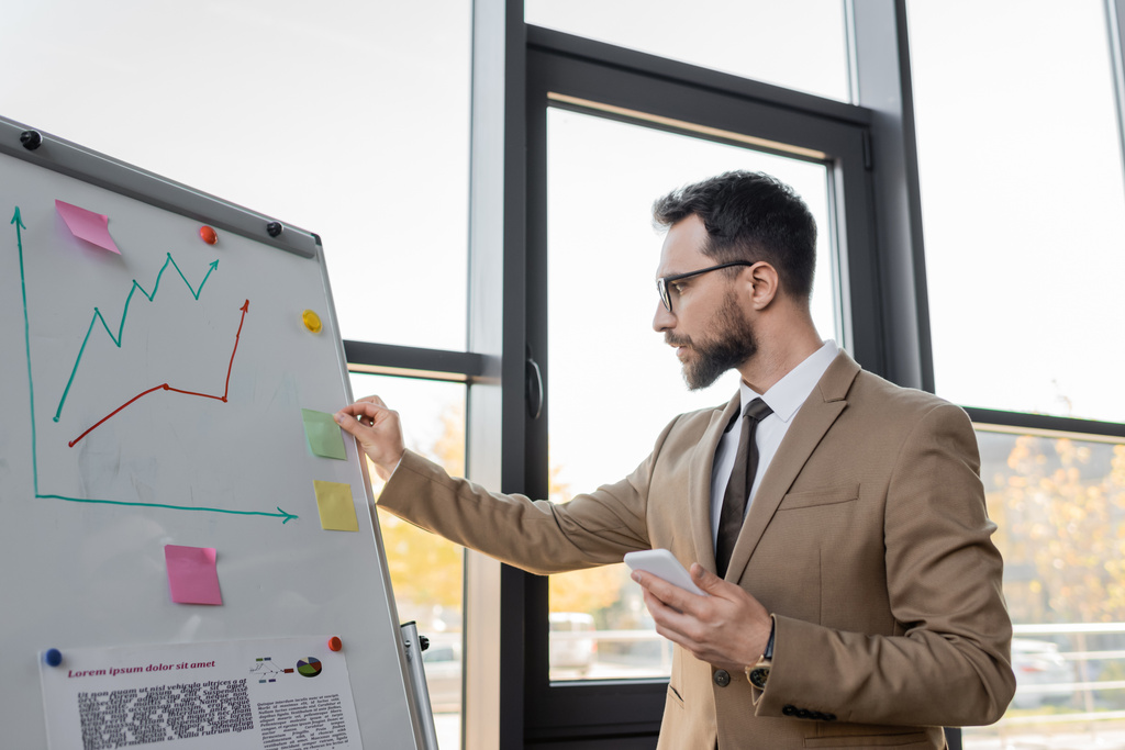 concentrated bearded corporate manager in stylish blazer and eyeglasses holding smartphone and working near flip chart and large windows in modern office - Photo, Image