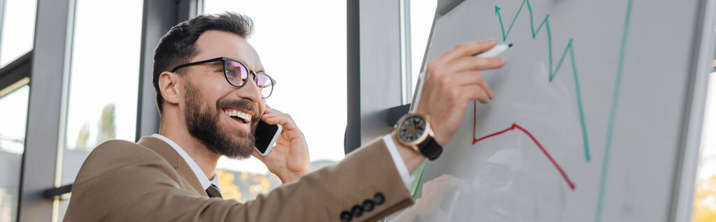 vzrušený vousatý podnikatel v brýlích, béžový blejzr a náramkové hodinky mluví na smartphonu, drží značku a při pohledu na rostoucí infografii na flip chart v kanceláři, banner - Fotografie, Obrázek