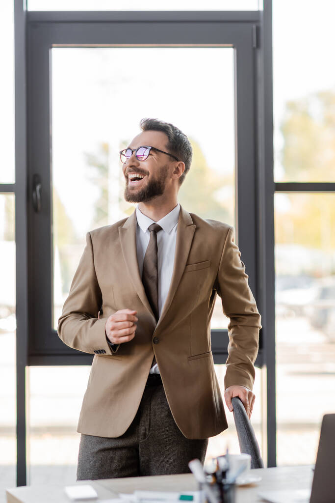hombre de negocios satisfecho y muy alegre en blazer beige de moda, corbata y anteojos mirando hacia otro lado cerca borrosa portátil, teléfono inteligente y taza de café en el escritorio en la oficina moderna - Foto, Imagen