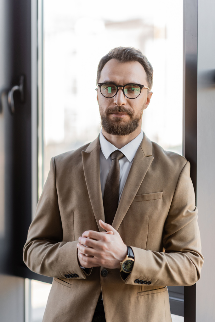 ambicioso empresário barbudo em blazer bege, óculos elegantes e relógio de pulso de luxo olhando para a câmera enquanto estava de pé com as mãos apertadas perto da janela no escritório - Foto, Imagem