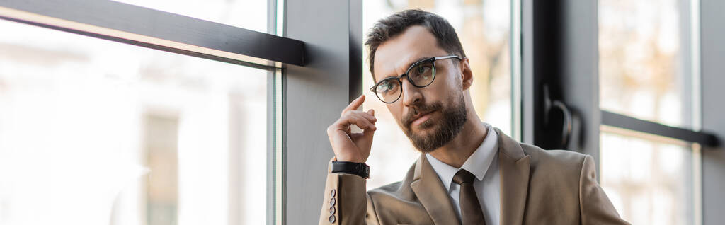 γενειοφόρος επιχειρηματίας με μοντέρνα γυαλιά, μπεζ σακάκι και γραβάτα κρατώντας το χέρι κοντά στο πρόσωπο, κοιτάζοντας μακριά και τη σκέψη κοντά σε παράθυρα στο σύγχρονο γραφείο, πανό - Φωτογραφία, εικόνα