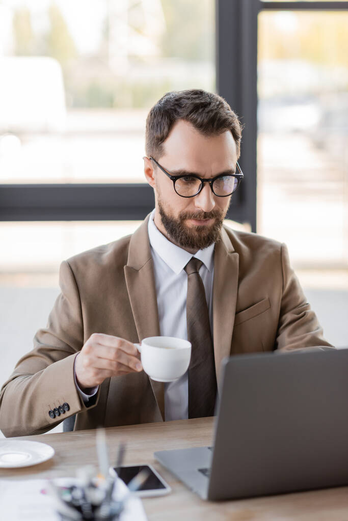 focused bearded businessman in stylish blazer, eyeglasses and tie holding coffee cup near mobile phone with blank screen and working on blurred laptop in office - Photo, Image