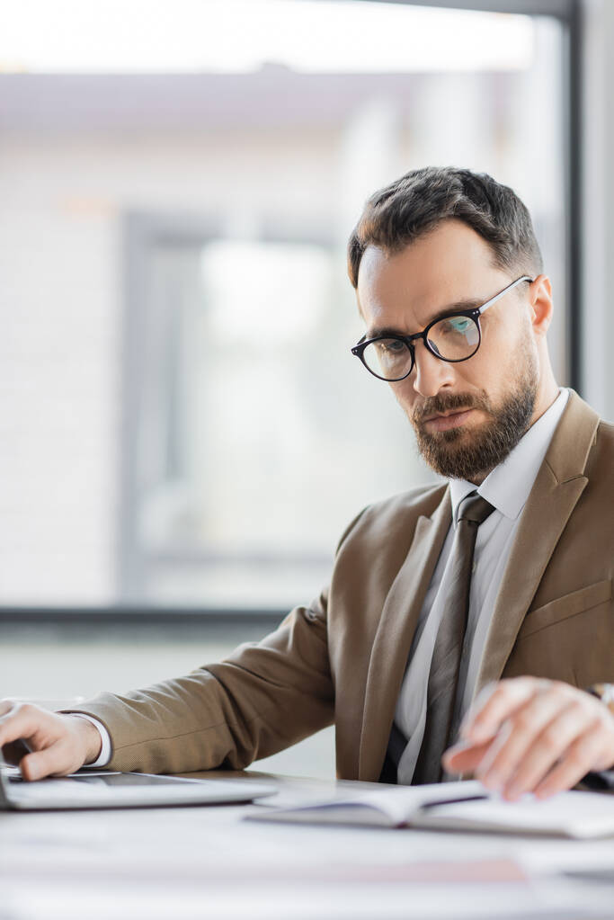 концентрований корпоративний менеджер з бородою і окулярами, одягнений в модний блейзер і краватку, дивиться на розмитий блокнот і працює на комп'ютері в офісі
 - Фото, зображення