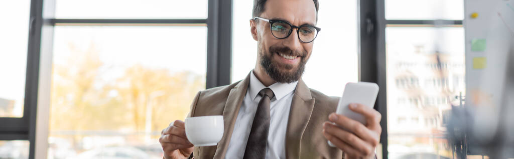 iloinen parrakas liikemies trendikkäässä bleiserissä, solmiossa ja silmälaseissa pitämässä kahvikuppia ja katselemassa matkapuhelinta toimistossa hämärtyneessä etualalla, banneri - Valokuva, kuva