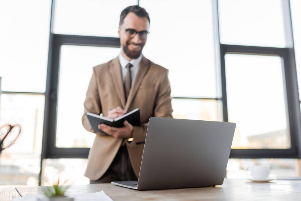 θετική γενειοφόρος επιχειρηματίας σε γυαλιά ηλίου, μπεζ σακάκι και γραβάτα στέκεται κοντά laptop και φλιτζάνι καφέ στο γραφείο εργασίας και γράφοντας στο σημειωματάριο σε θολή φόντο - Φωτογραφία, εικόνα