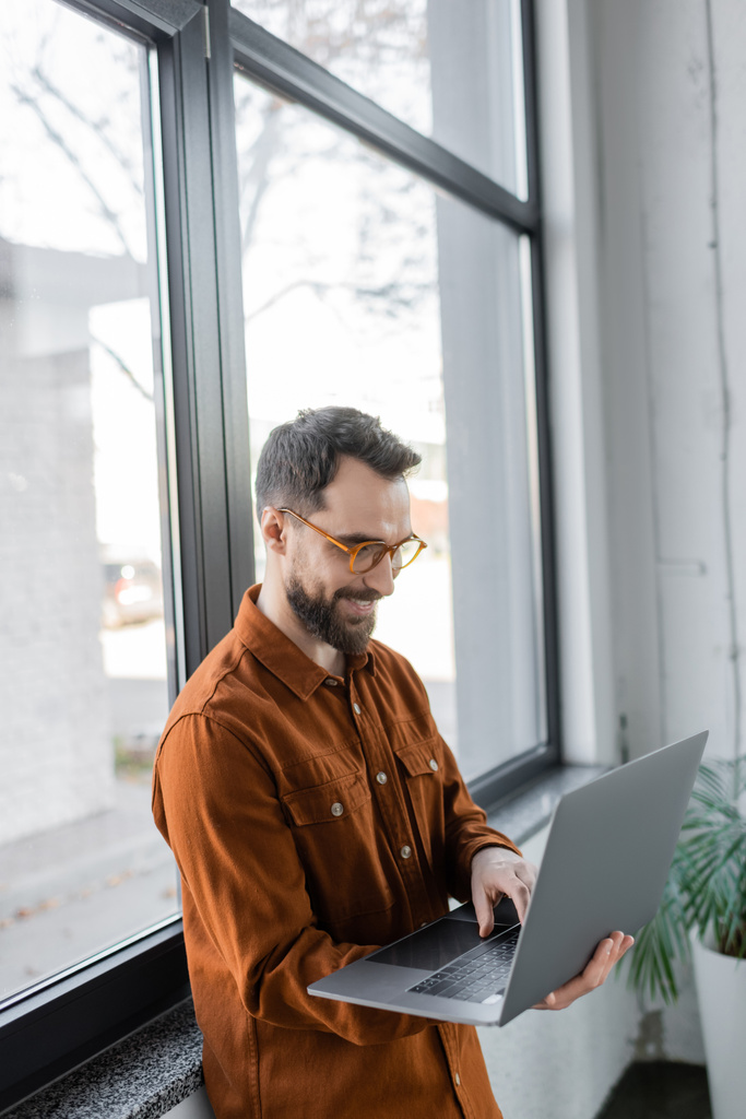 positiver und erfolgreicher bärtiger Unternehmer mit trendiger Brille und stylischem Hemd, der neben großen Fenstern im modernen Büro steht und am Laptop arbeitet - Foto, Bild