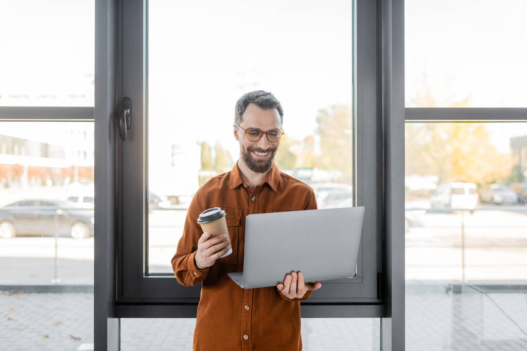 homem de negócios satisfeito e barbudo em óculos e camisa elegante sorrindo enquanto estava de pé com bebida takeaway e laptop perto de janelas no escritório contemporâneo - Foto, Imagem