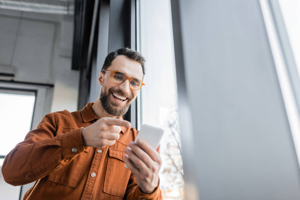 low angle view of excited entrepreneur with beard and eyeglasses, wearing stylish shirt, pointing with finger at mobile phone during video call in modern office  - Photo, Image