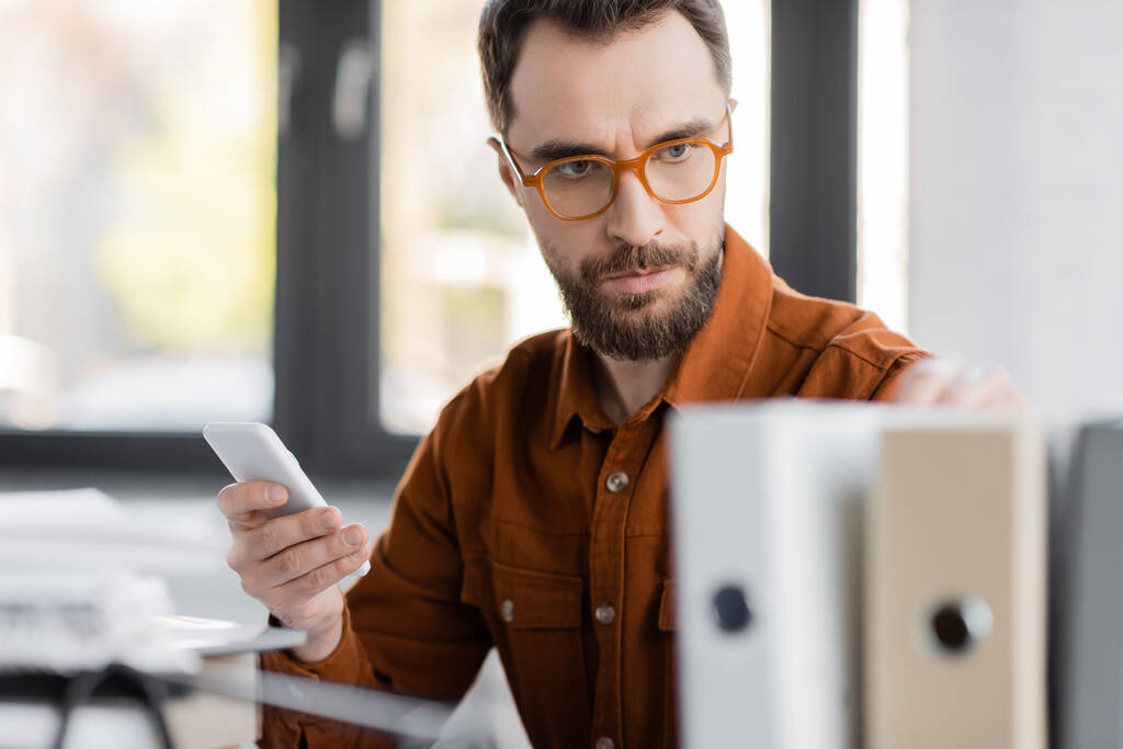 концентрований і серйозний бізнесмен в стильних окулярах і сорочках тримає мобільний телефон і дивиться на розмиті папки під час роботи над продуктивністю бізнесу в офісі
 - Фото, зображення