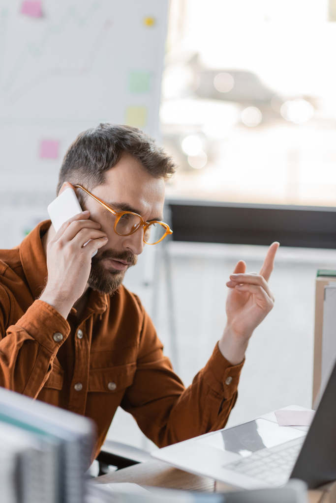 εστιασμένος γενειοφόρος επιχειρηματίας με γυαλιά ηλίου και πουκάμισο που μιλάει σε smartphone και δείχνει χειρονομία προσοχής κοντά σε φορητό υπολογιστή και θολά σημειωματάρια στο χώρο εργασίας στο γραφείο - Φωτογραφία, εικόνα